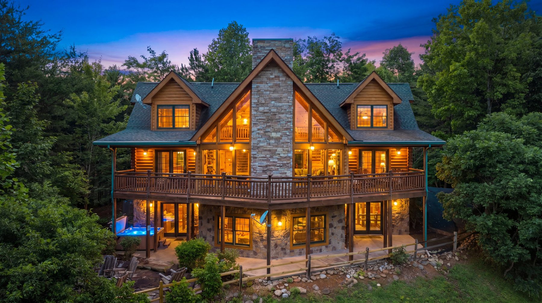 A Colonial Properties log cabin lit up at dusk in the Smoky Mountains, with a wraparound deck, stone chimney and hot tub