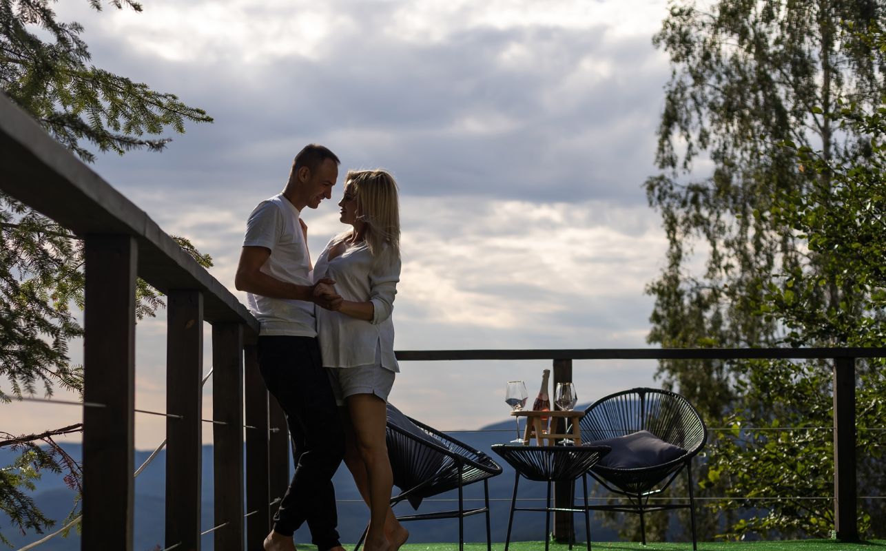 7 Features That Make Our Smoky Mountain Honeymoon Cabins the Perfect Escape for Two Romantic couple sharing a quiet moment on a scenic mountain-view deck with modern outdoor seating, wine glasses, and a bottle of rosé at sunset.